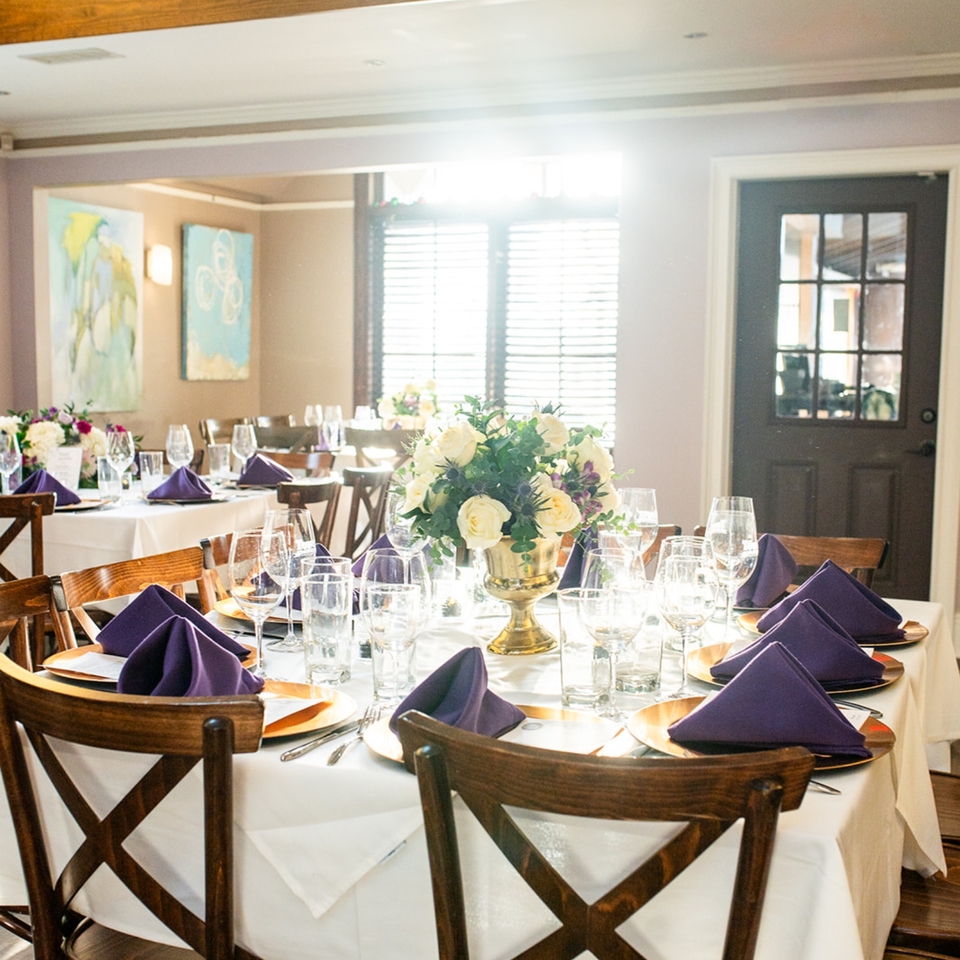 Elegant restaurant dining room with white tablecloths, purple folded napkins, glassware, floral centerpieces, and wooden chairs.