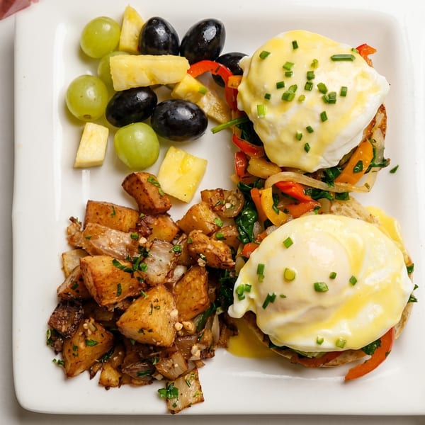 Eggs Benedict with hollandaise sauce, home fries, and a side of fruit on a white plate.