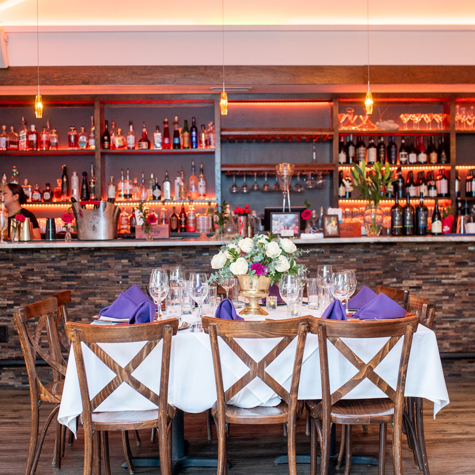 Elegant restaurant dining table with white linens, purple napkins, floral centerpiece, wooden chairs, and illuminated bar stocked with bottles.