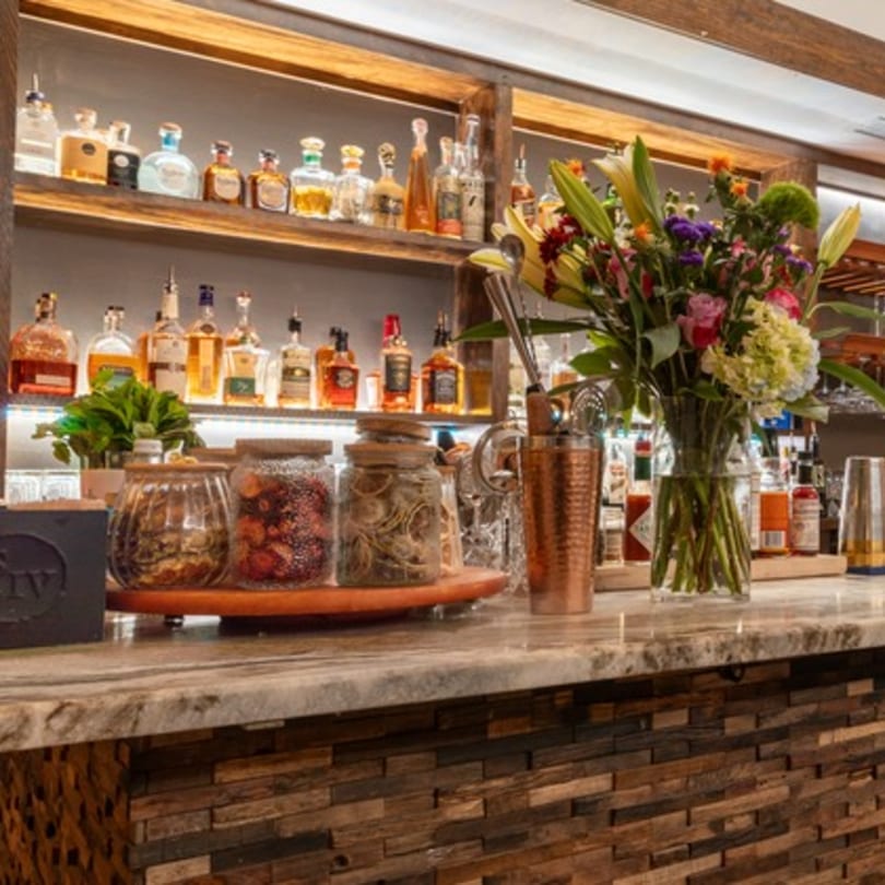 A well-stocked bar with liquor bottles on shelves, a floral arrangement, and cocktail ingredients.
