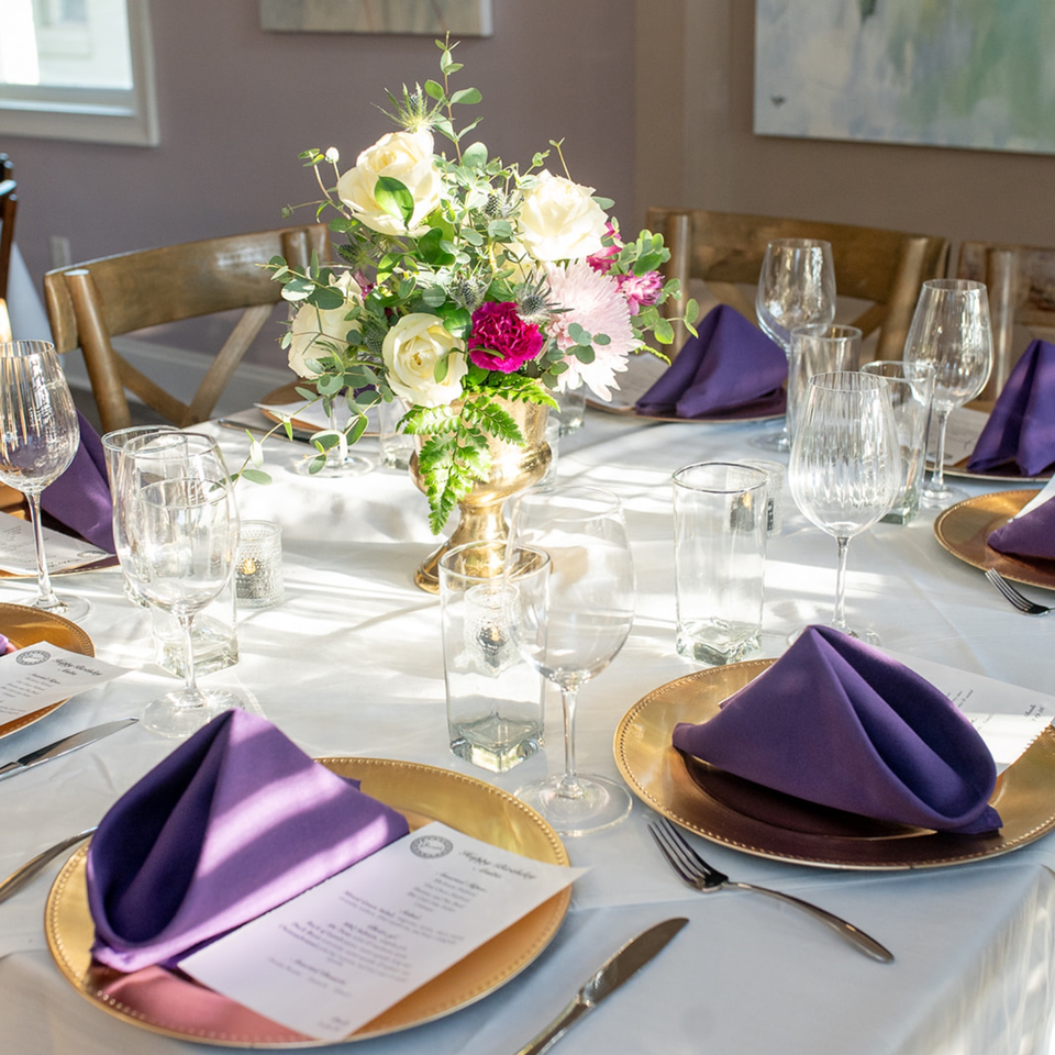 Elegant restaurant table set with gold chargers, purple napkins, glassware, menus, and a colorful floral centerpiece.