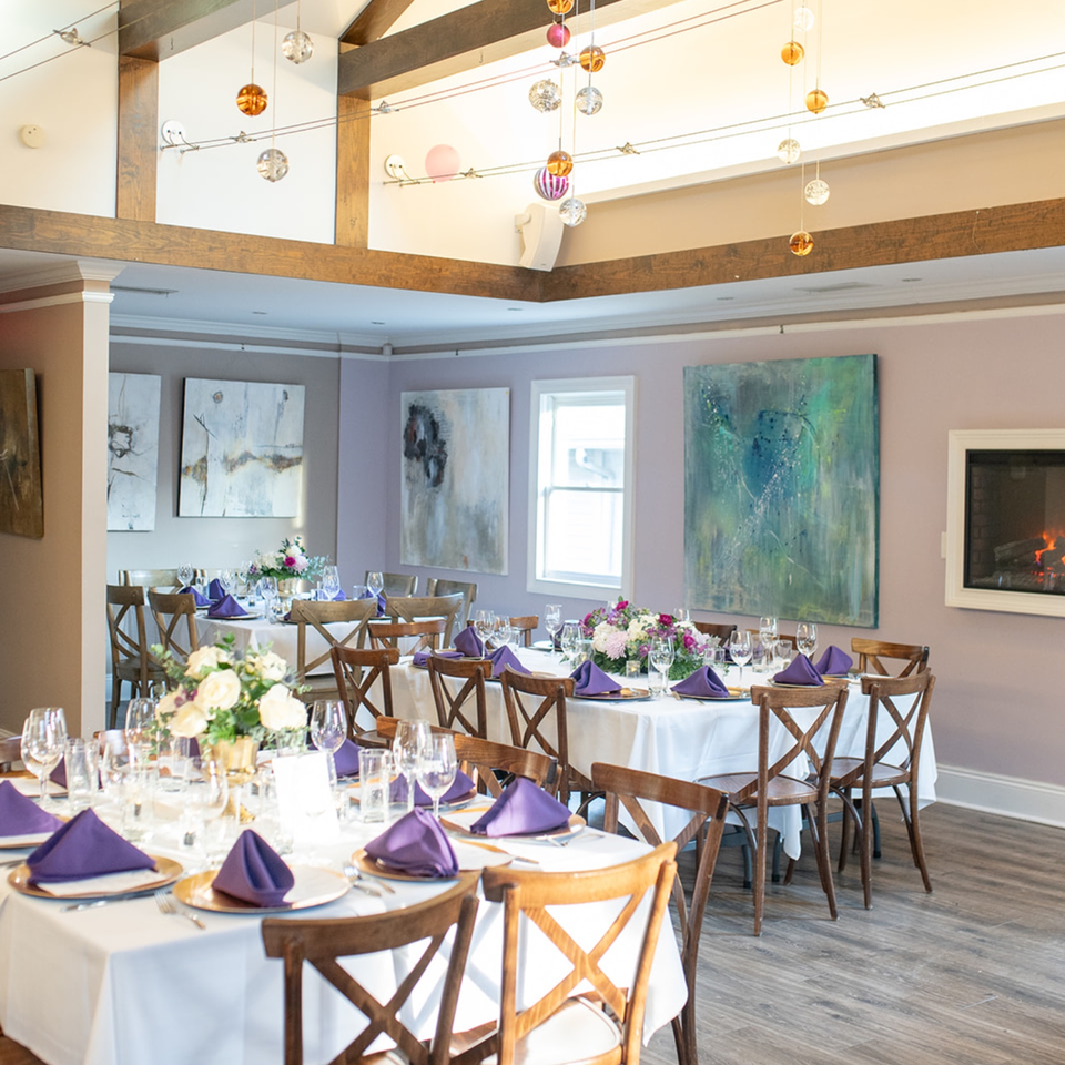 Elegant restaurant dining room with white tablecloths, purple napkins, floral centerpieces, artwork, and a glowing fireplace.