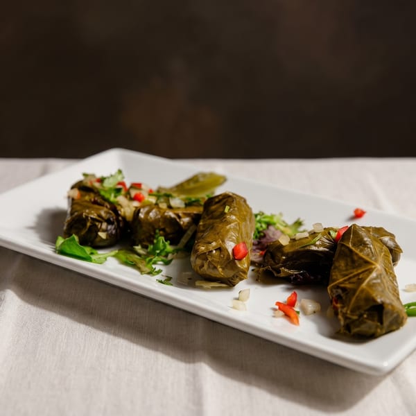 A white rectangular plate holds several dolmades, garnished with chopped herbs, red pepper, and onion.