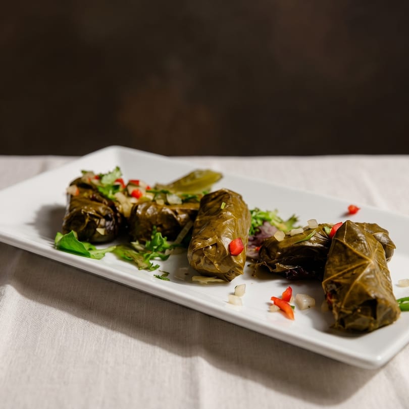 A white rectangular plate holds several dolmades, garnished with chopped herbs, red pepper, and onion.
