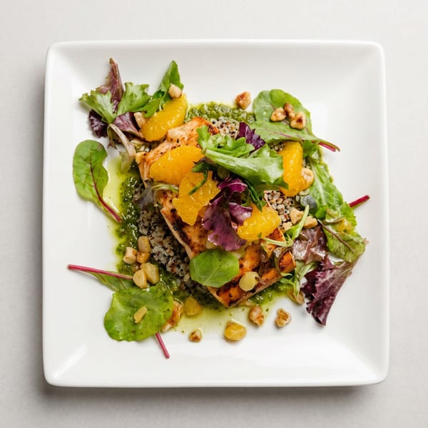Grilled salmon with quinoa, orange segments, mixed greens, and a green sauce on a white plate.